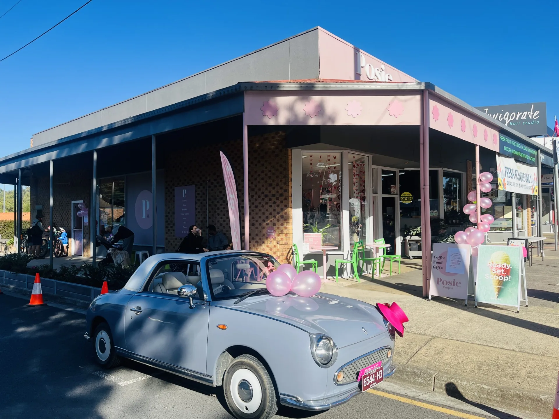 The Posie Project storefront on Mount Dandenong Tourist Road in Montrose — pink awning with cherry-blossom cutouts, pastel chairs out the front, and a vintage blue Figaro parked at the kerb